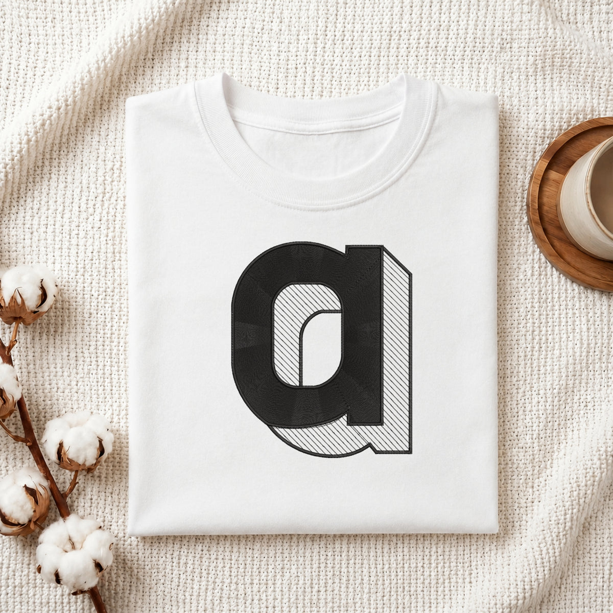 A neatly folded white crewneck t-shirt featuring a stitched 3D block lowercase letter "a" design, styled with cotton branches and a wooden coaster.