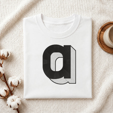 A neatly folded white crewneck t-shirt featuring a stitched 3D block lowercase letter "a" design, styled with cotton branches and a wooden coaster.