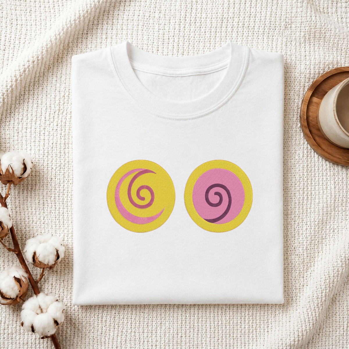 A folded white T-shirt laid flat on a textured cream fabric background, featuring two circular graphic prints on the chest: one yellow circle with a pink spiral and one pink circle with a yellow spiral. Cotton bolls and part of a wooden cup are visible as styled props.