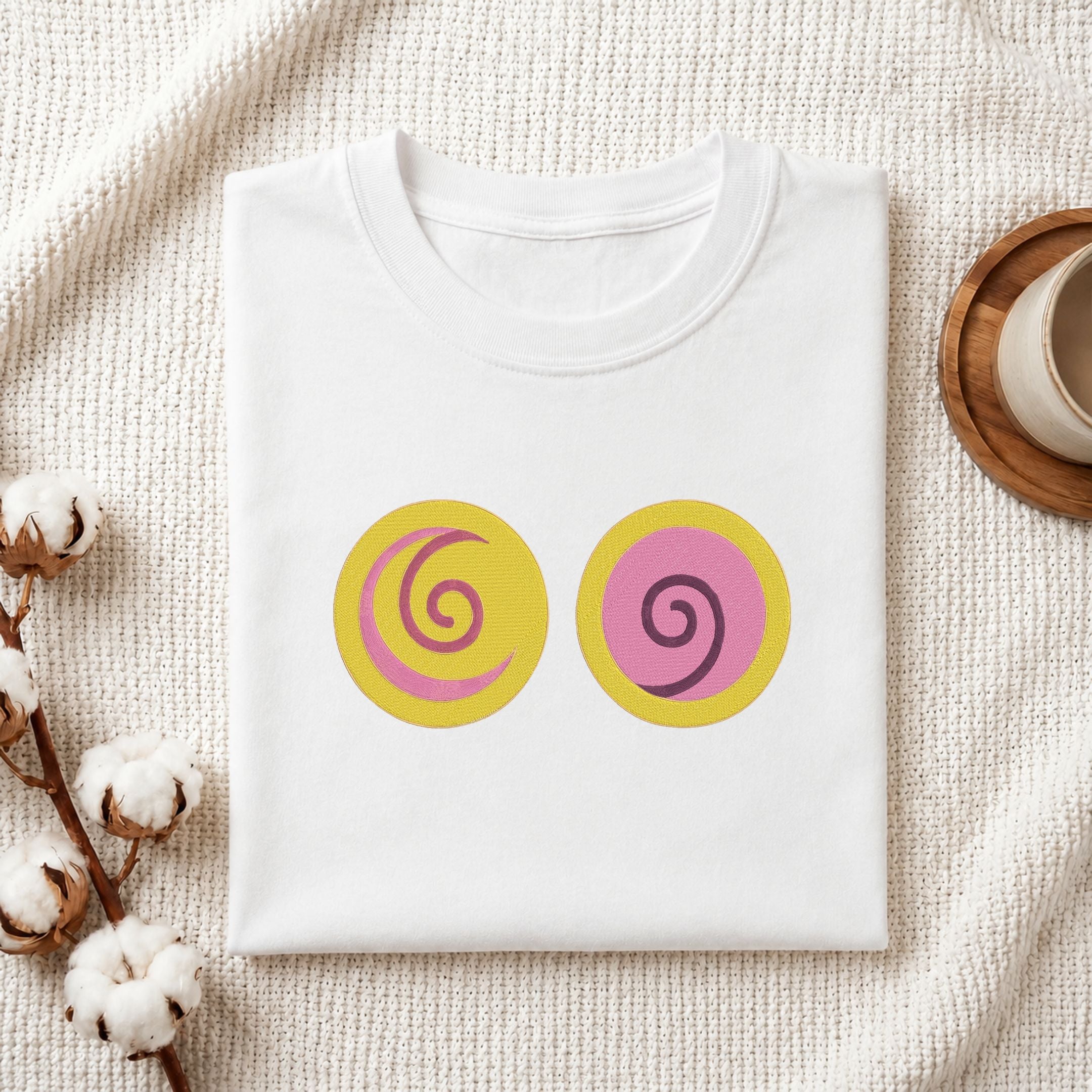 A folded white T-shirt laid flat on a textured cream fabric background, featuring two circular graphic prints on the chest: one yellow circle with a pink spiral and one pink circle with a yellow spiral. Cotton bolls and part of a wooden cup are visible as styled props.