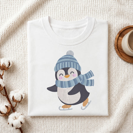 A folded white crewneck t-shirt featuring a cute cartoon penguin wearing a blue striped knit hat and matching scarf, skating on ice. The shirt is styled in a cozy flat-lay setting on a textured cream fabric, with cotton bolls and a wooden cup in the background.