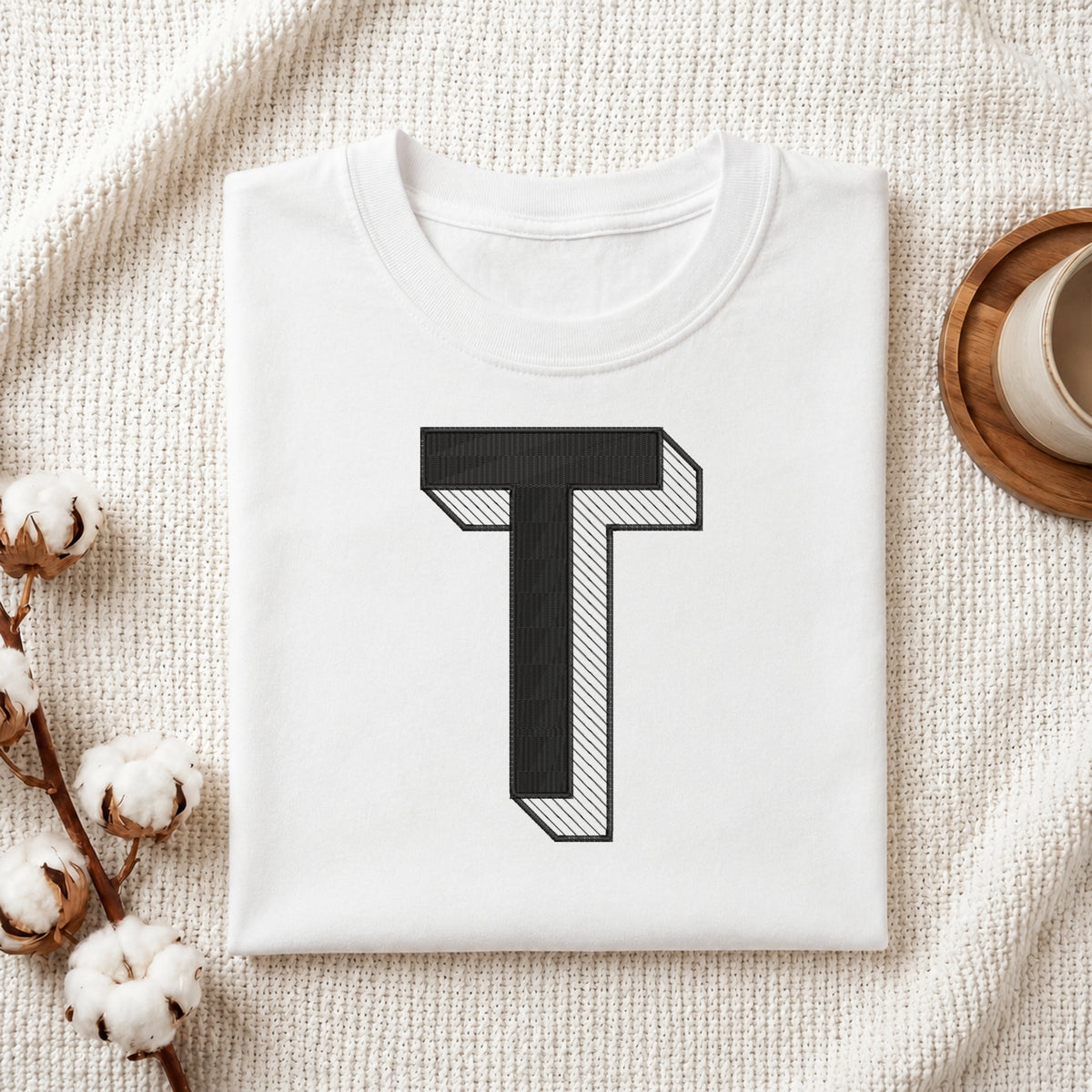 A neatly folded white crewneck t-shirt featuring a stitched 3D block letter "T" design, styled with cotton branches and a wooden coaster.