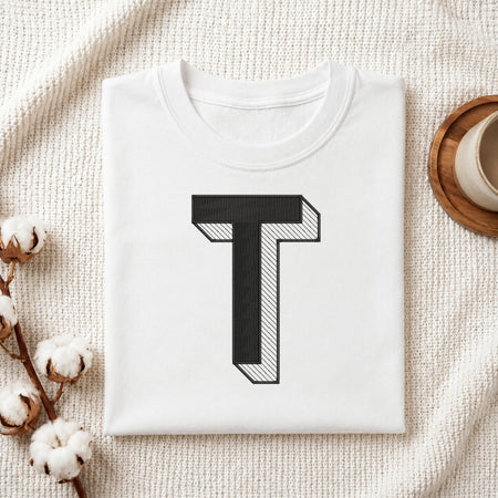 A neatly folded white crewneck t-shirt featuring a stitched 3D block letter "T" design, styled with cotton branches and a wooden coaster.