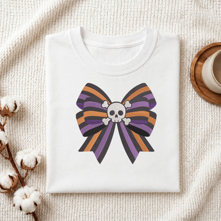 A neatly folded white t-shirt featuring the embroidered striped skull bow design, displayed on a textured white knit background with cotton stems and a wooden cup.