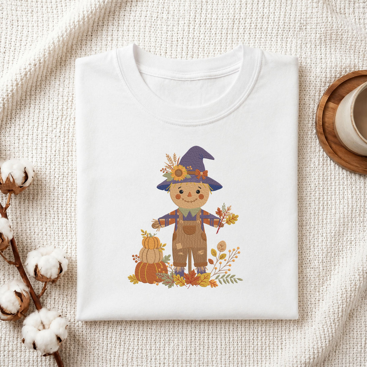 Folded white cotton t-shirt mockup featuring a cute autumn scarecrow machine embroidery design, styled with cotton stems and a wooden cup