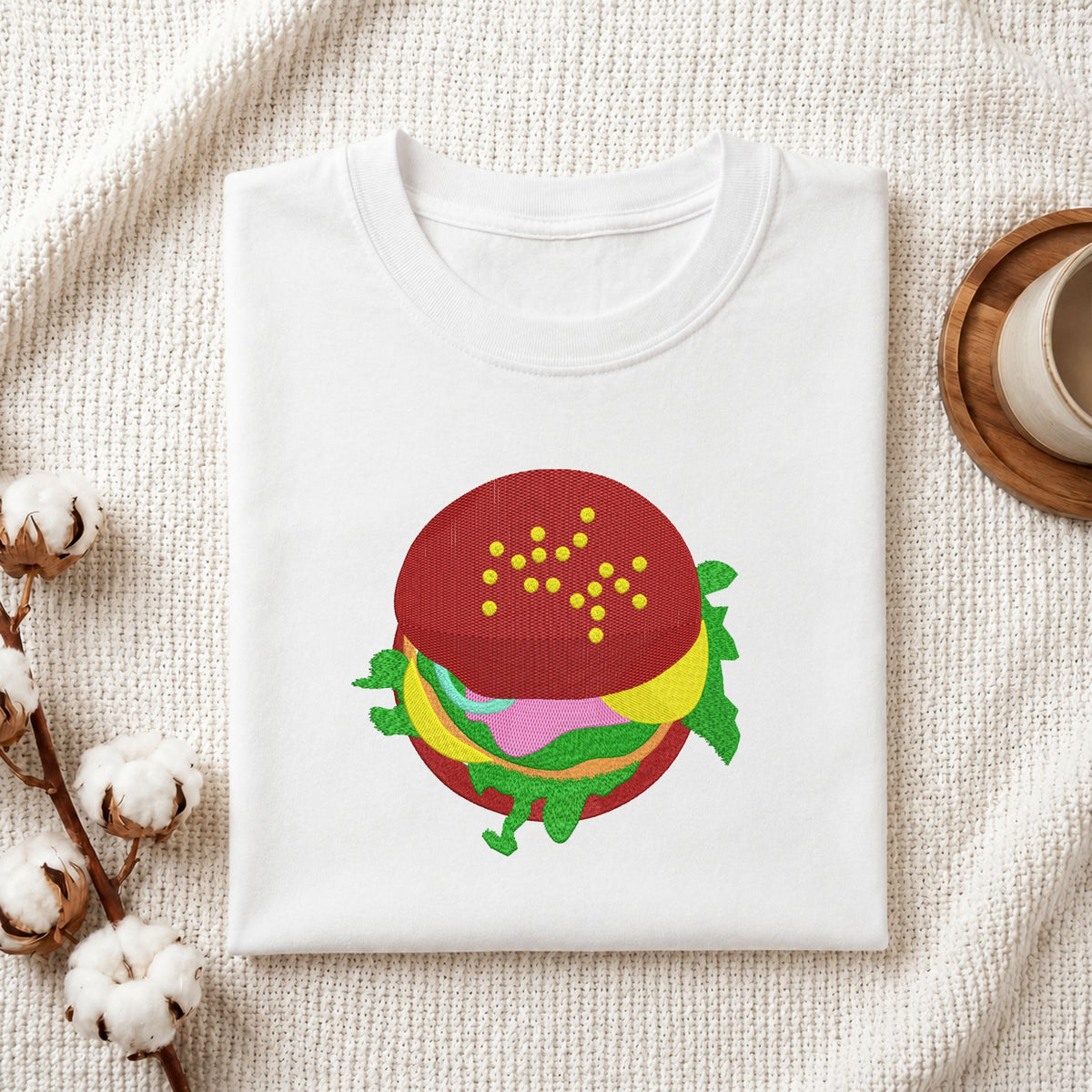 Folded white cotton t-shirt mockup featuring a delicious burger machine embroidery design, styled with cotton stems and a wooden cup   