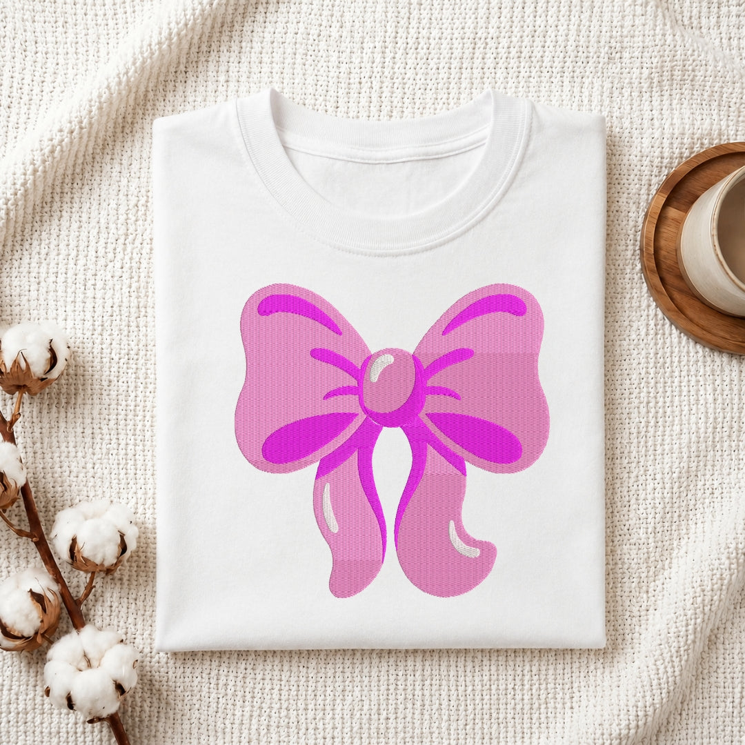 A folded white crewneck t-shirt featuring a large pink bow graphic on the front, styled on a textured cream fabric with cotton bolls and a wooden cup in the background.