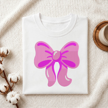 A folded white crewneck t-shirt featuring a large pink bow graphic on the front, styled on a textured cream fabric with cotton bolls and a wooden cup in the background.