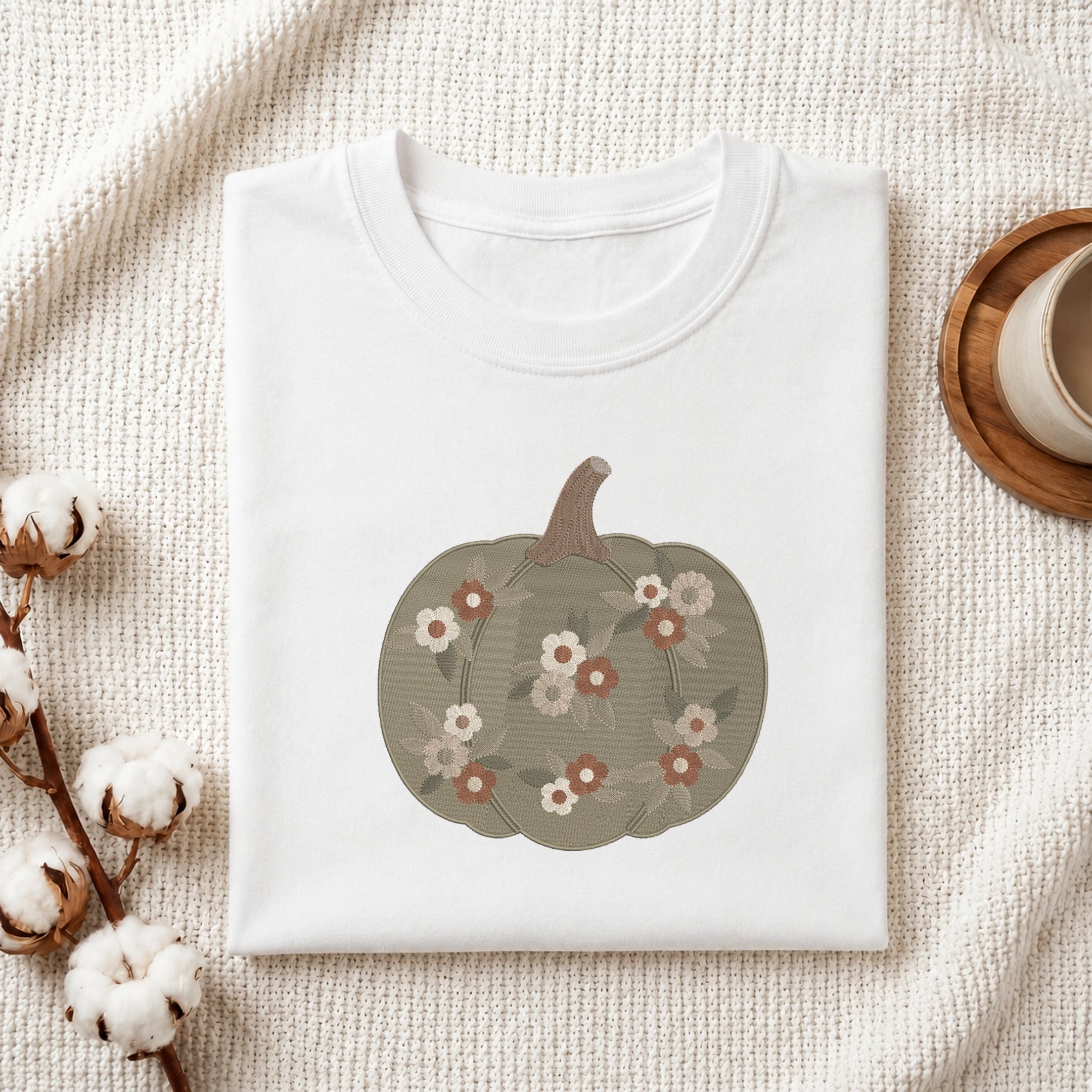 A folded white crewneck t-shirt with a graphic of a muted green pumpkin decorated with small white, pink, and brown flowers. The shirt is styled in a flat-lay on a textured cream knit blanket, with cotton bolls on the left and a wooden cup on the right.