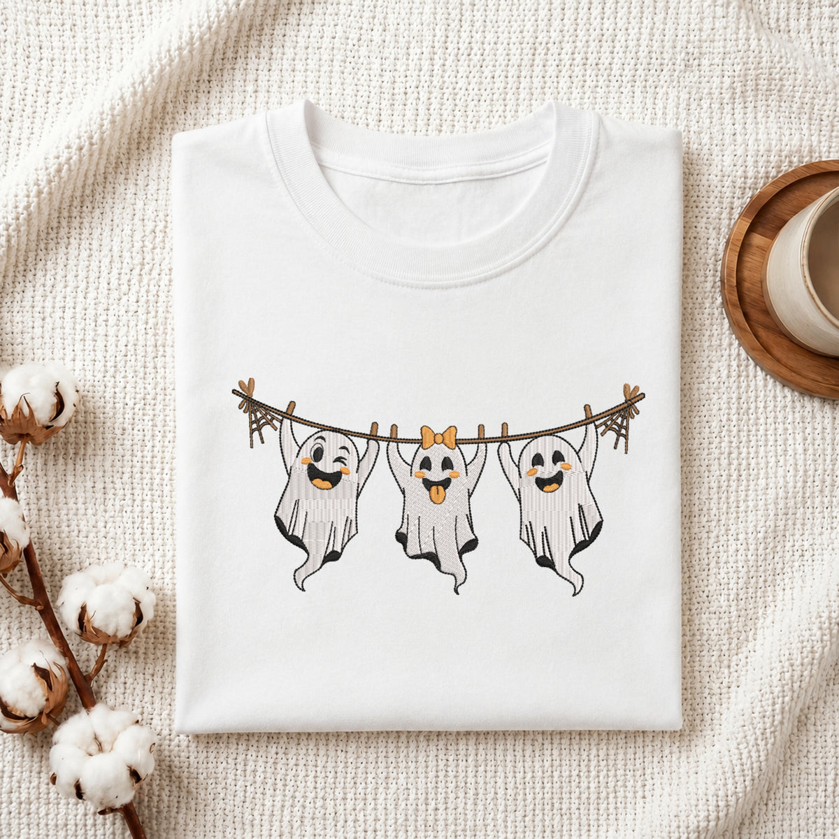 A folded white crewneck t-shirt with a graphic of three smiling ghosts hanging from a clothesline, styled on a textured cream knit blanket with cotton bolls and a wooden cup in the background.