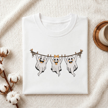 A folded white crewneck t-shirt with a graphic of three smiling ghosts hanging from a clothesline, styled on a textured cream knit blanket with cotton bolls and a wooden cup in the background.