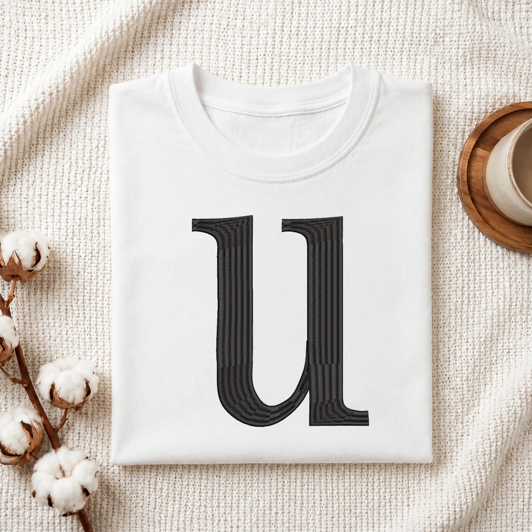 A folded white crewneck t-shirt featuring a large, stylized black letter 'U' printed on the front, styled in a flat lay on a textured cream fabric with cotton bolls and a wooden cup.
