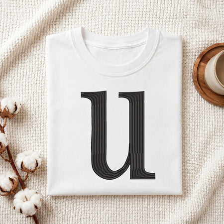 A folded white crewneck t-shirt featuring a large, stylized black letter 'U' printed on the front, styled in a flat lay on a textured cream fabric with cotton bolls and a wooden cup.