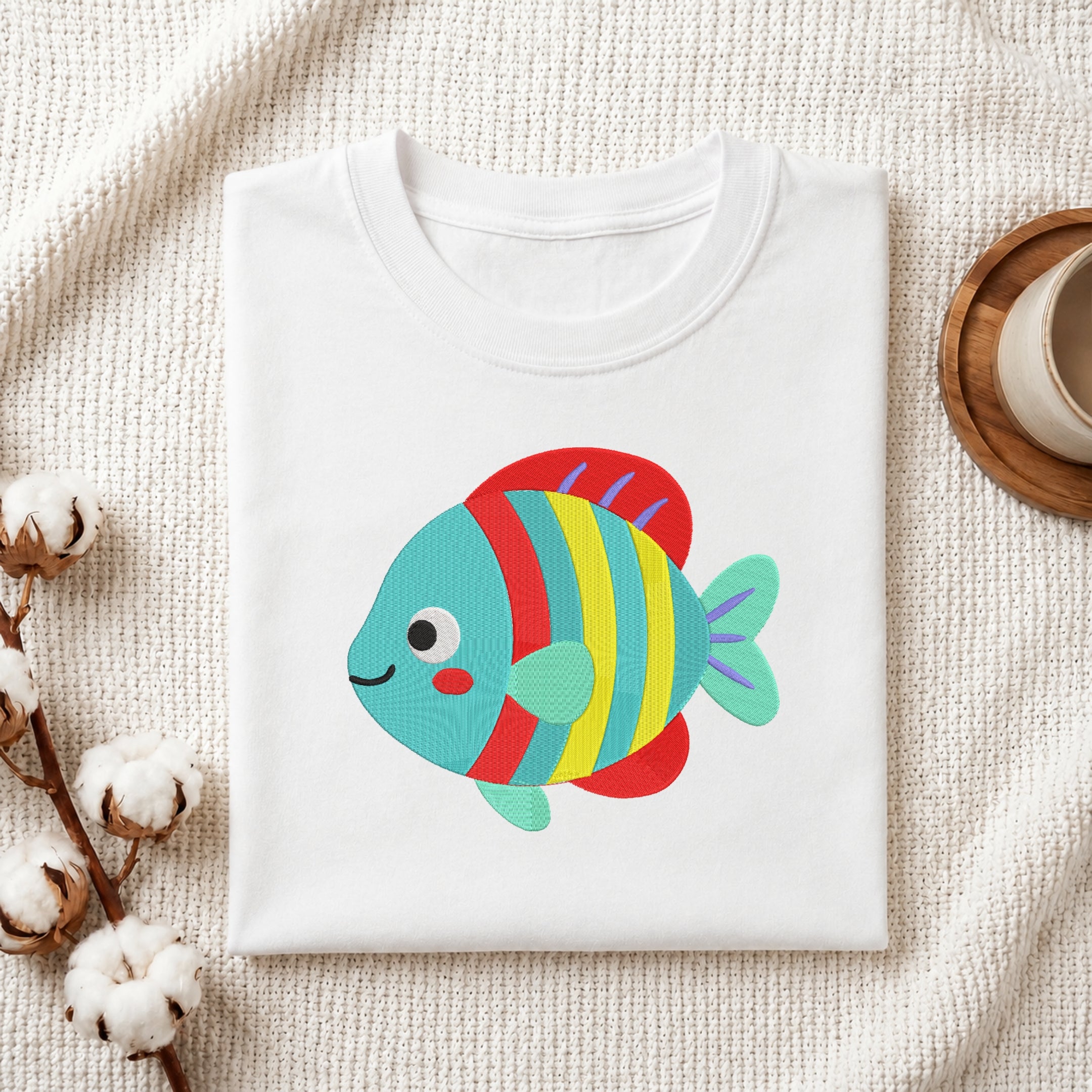 A folded white crewneck t-shirt featuring a colorful embroidered cartoon fish with red, yellow, and blue stripes and a teal body, styled on a textured cream fabric with cotton bolls and a wooden cup.