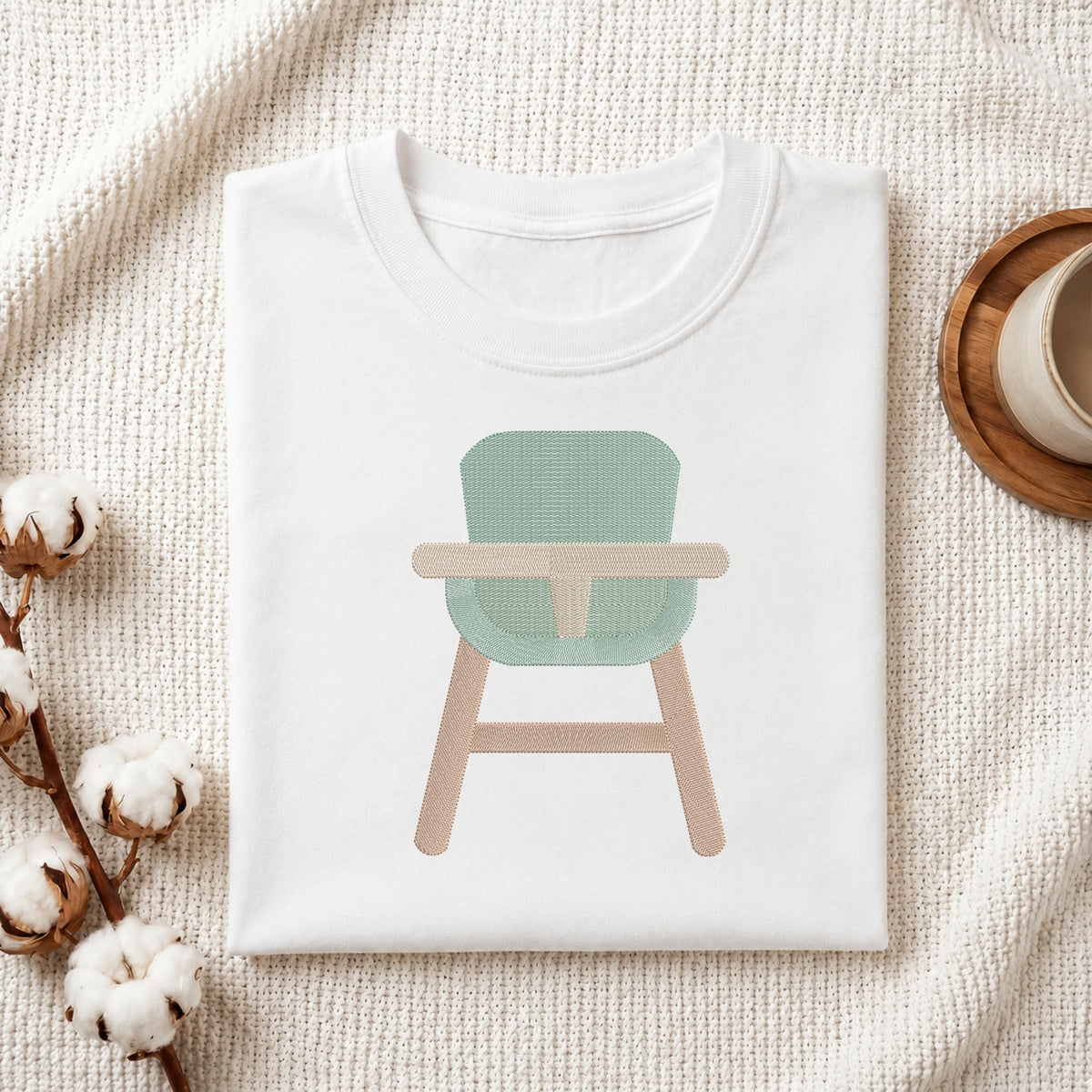 A folded white T-shirt laid flat on a textured cream fabric background, featuring a printed illustration of a high chair with a mint green seat and light wood legs. Cotton bolls and a small cup on a wooden saucer are placed around it as styled props.