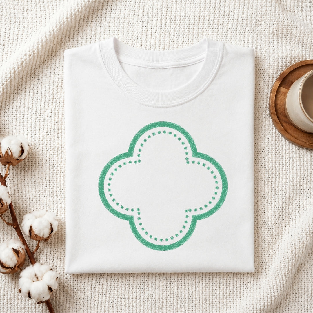 A folded white crewneck t-shirt with a green four-leaf clover graphic, styled on a textured cream blanket with cotton bolls and a wooden cup in the background.