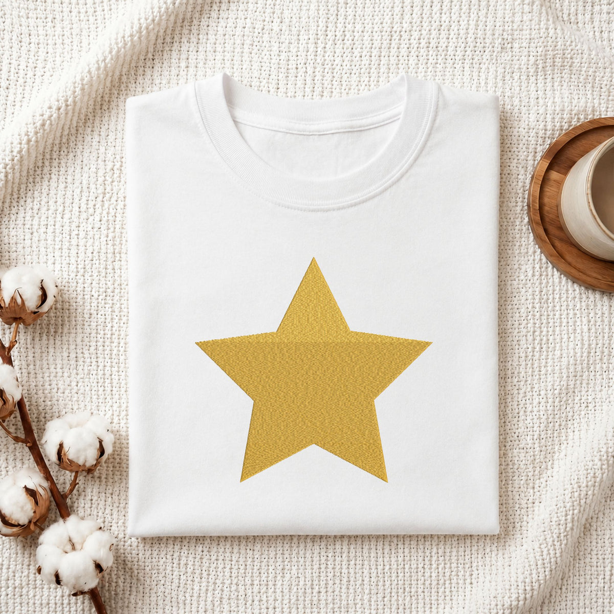 Folded white cotton t-shirt mockup featuring a golden star machine embroidery design, styled with cotton stems and a wooden cup