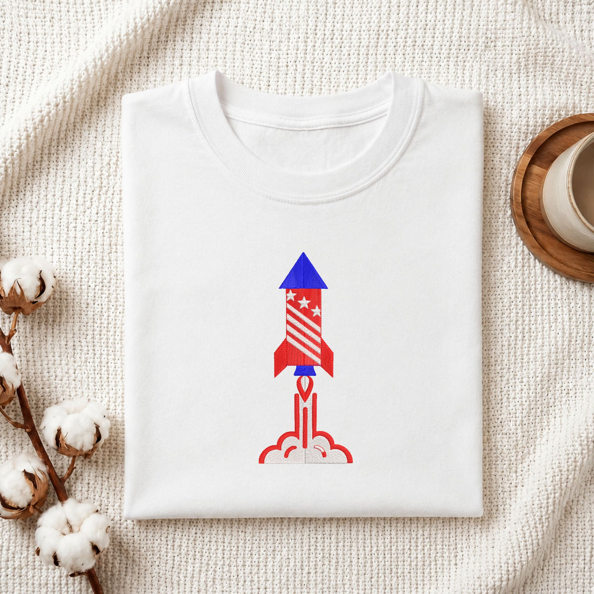 Folded white cotton t-shirt mockup featuring a patriotic space rocket machine embroidery design, styled with cotton stems and a wooden cup.