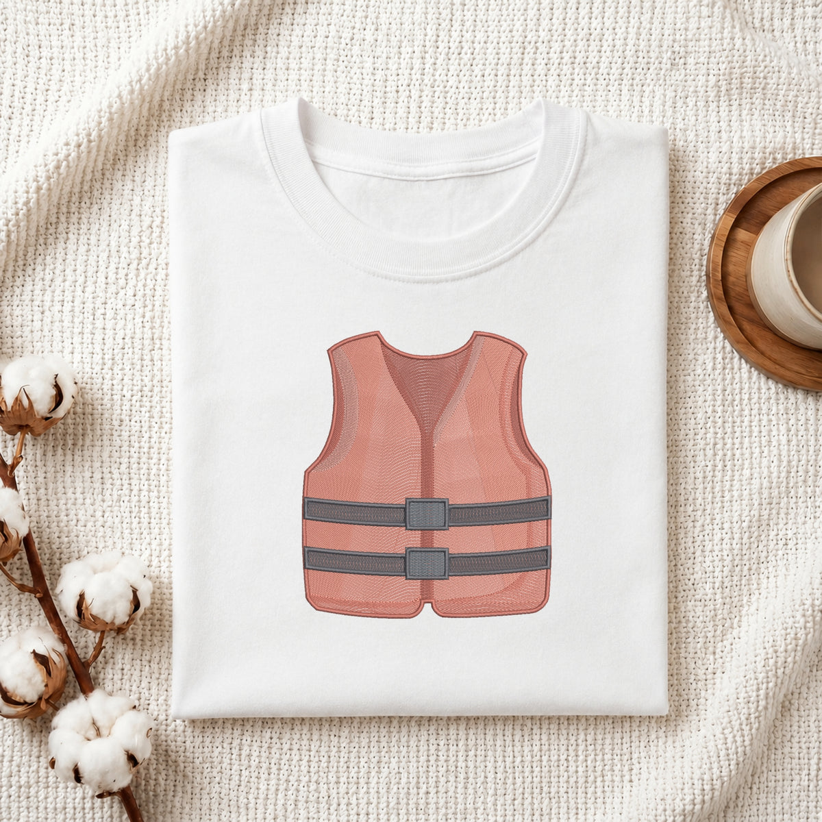 A folded white T-shirt laid flat on a textured cream fabric background, featuring a printed illustration of a pink life vest with gray straps on the front. Decorative cotton bolls and a small wooden cup appear at the edges.