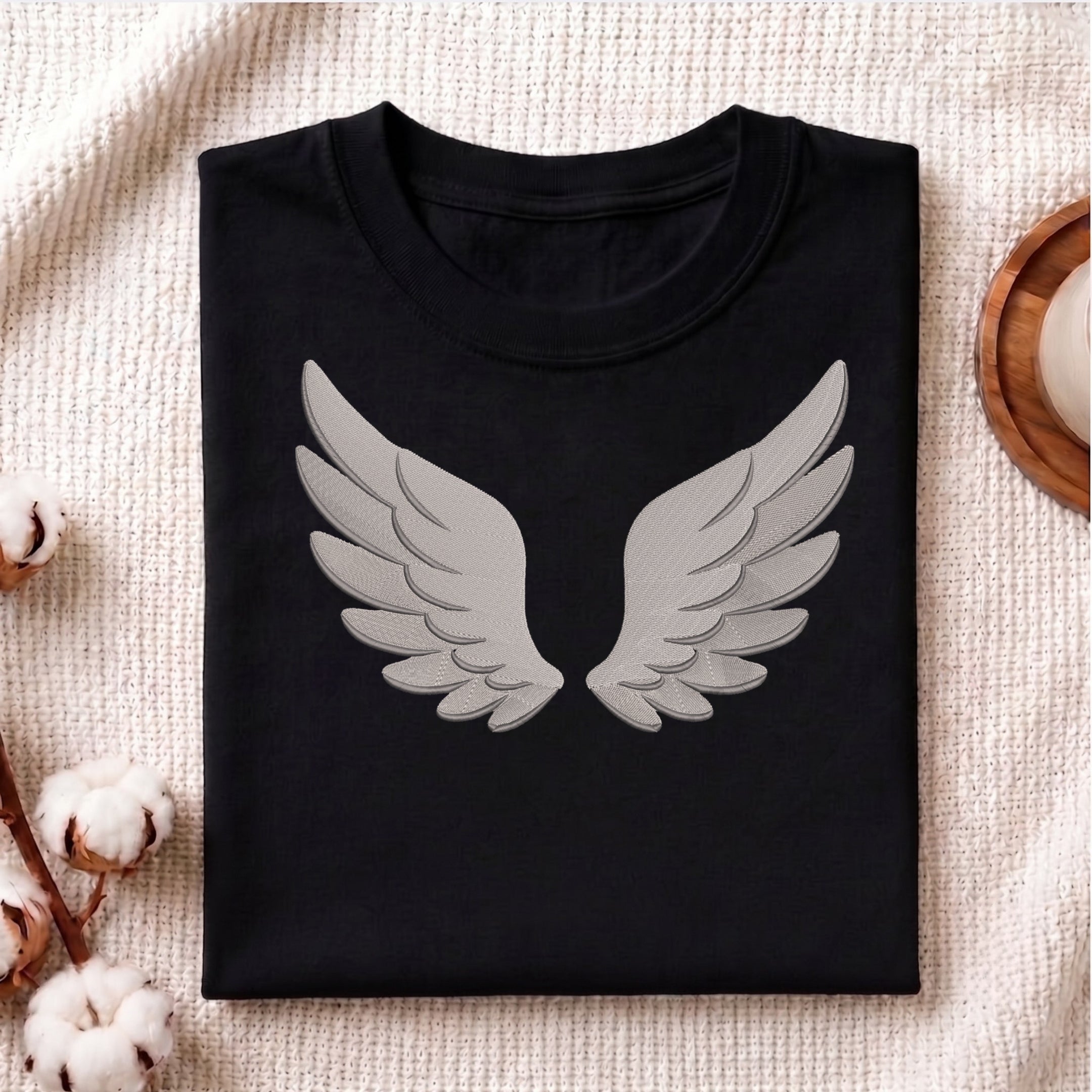 A folded black crewneck t-shirt featuring a large gray embroidered angel wings graphic on the front, styled on a textured beige blanket with cotton bolls and a wooden tray.