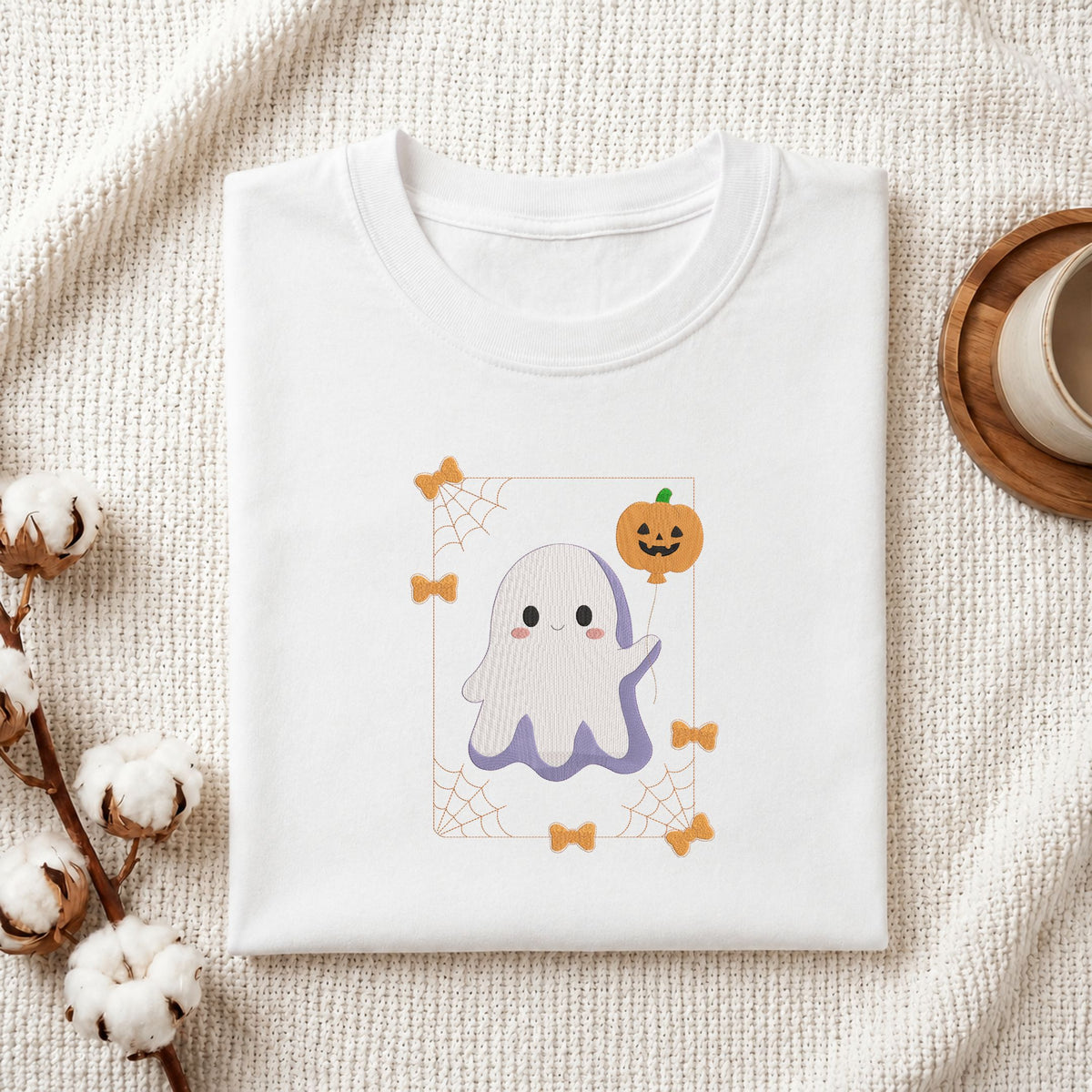 Folded white cotton t-shirt mockup featuring a cute ghost machine embroidery design, styled with cotton stems and a wooden cup