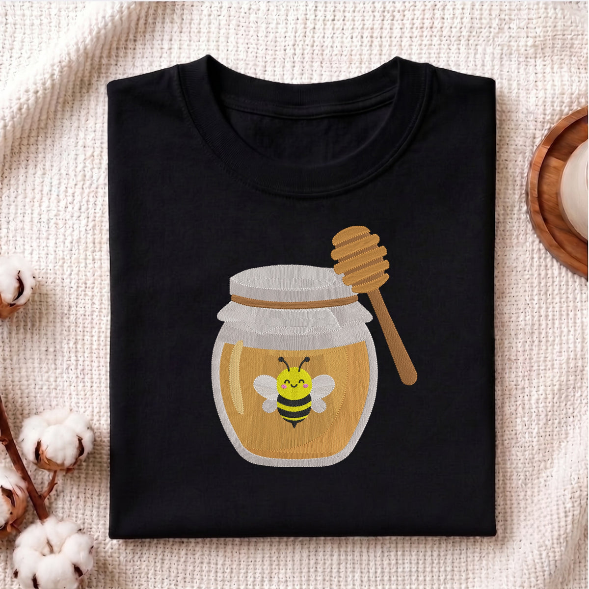 A folded black t-shirt with a graphic of a honey jar and a smiling bee, styled on a textured beige fabric with cotton bolls and a wooden spoon in the background.