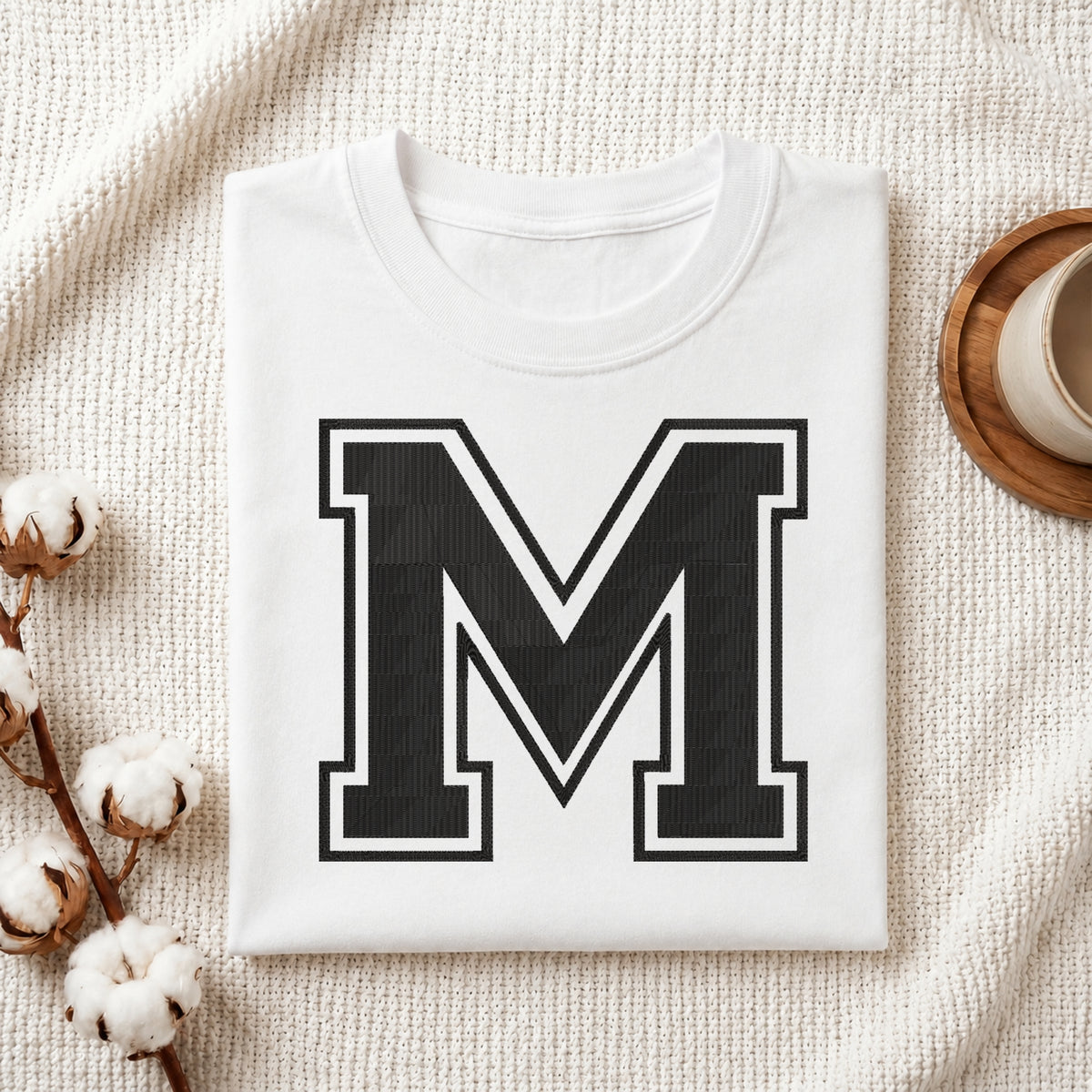 A folded white crewneck t-shirt with a large black block letter 'M' outlined in white on the front, styled on a textured cream knit blanket with cotton bolls and a small cup in the background.