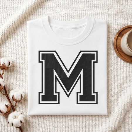 A folded white crewneck t-shirt with a large black block letter 'M' outlined in white on the front, styled on a textured cream knit blanket with cotton bolls and a small cup in the background.