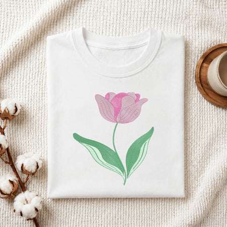 A folded white crewneck t-shirt featuring a pink tulip with green leaves printed on the front, styled on a textured cream fabric with cotton bolls and a wooden cup in the background.