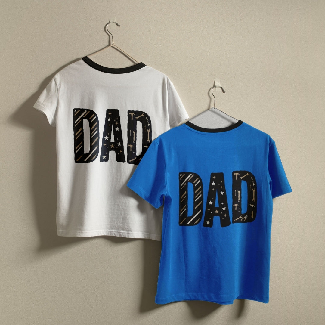 Two ringer-style short-sleeve T-shirts hanging on hangers against a plain beige wall. The back of one shirt is white with a black collar and a large black 'DAD' graphic, and the front of the other shirt is bright blue with a black collar and a large black 'DAD' graphic.