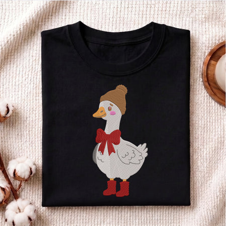 A folded black crewneck t-shirt featuring a cartoon graphic of a white goose wearing a brown beanie, red bow, and red boots, displayed on a textured beige fabric background with decorative cotton bolls.