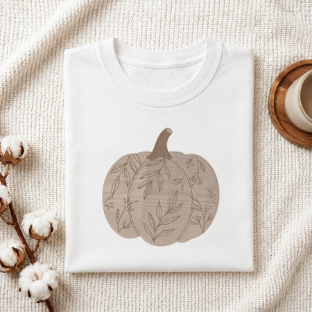 A neatly folded white crewneck t-shirt featuring a stitched floral pattern pumpkin design, styled with cotton branches and a wooden coaster.