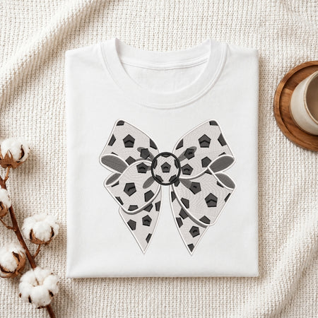 A folded white crewneck t-shirt with a large bow graphic printed on the front, styled on a textured cream fabric background with cotton bolls and a wooden cup.