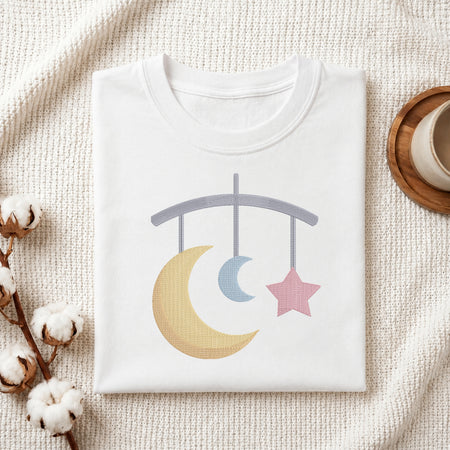A folded white baby t-shirt with a pastel-colored mobile graphic (yellow crescent moon, blue crescent moon, pink star) on the front, styled on a textured cream blanket with cotton bolls and a wooden cup.