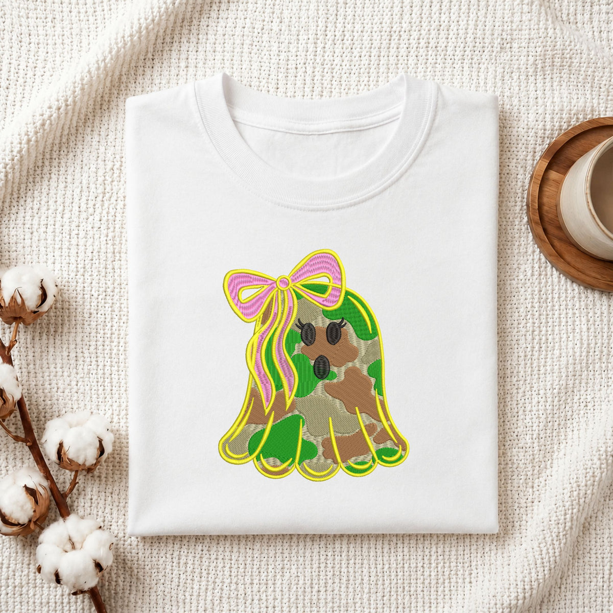 Folded white cotton t-shirt mockup featuring a cute camo ghost machine embroidery design, styled with cotton stems and a wooden cup   