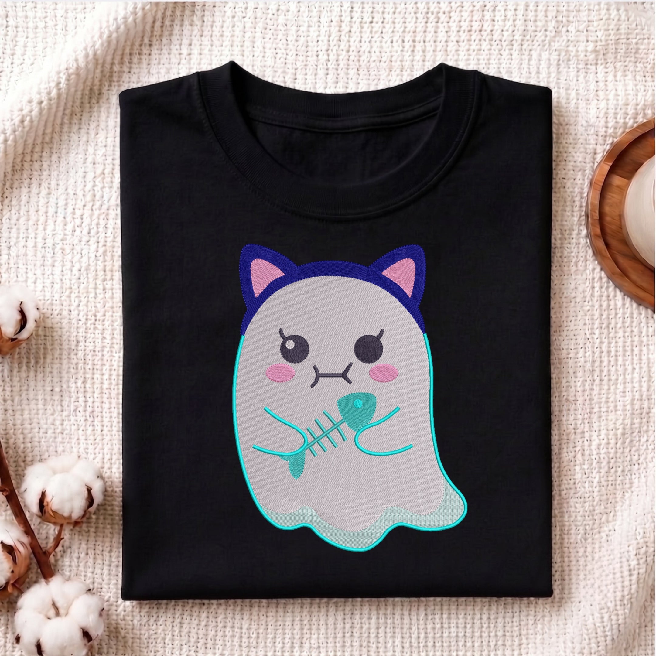 A folded black t-shirt with a cute cartoon ghost graphic holding a fish skeleton, displayed on a textured beige fabric background with decorative cotton bolls.