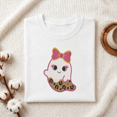 A folded white t-shirt with a cute cartoon ghost graphic, styled on a textured cream blanket with cotton bolls and a wooden cup in the background.