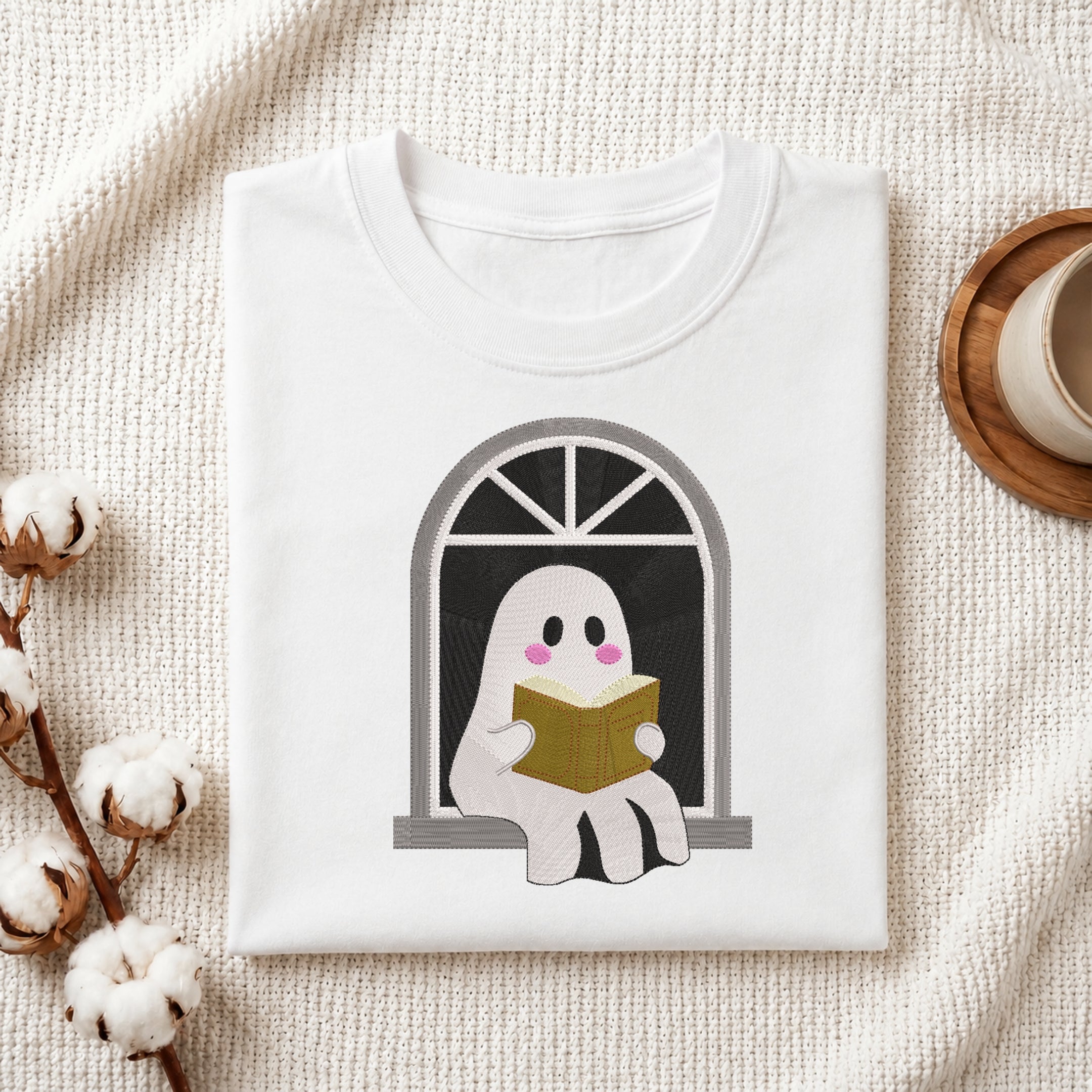 A folded white T-shirt with a cute cartoon ghost graphic sitting by a window and reading a book, styled on a textured cream fabric with cotton stems and a cup in the background.