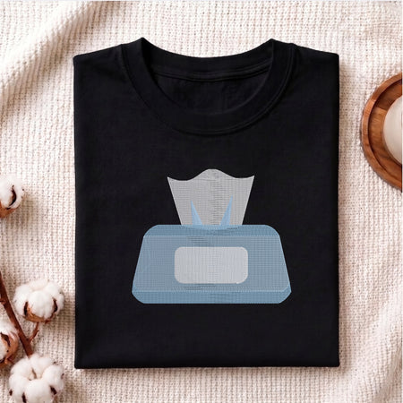 A neatly folded black crewneck t-shirt featuring a stitched blue tissue box design, styled with cotton branches and a wooden coaster.