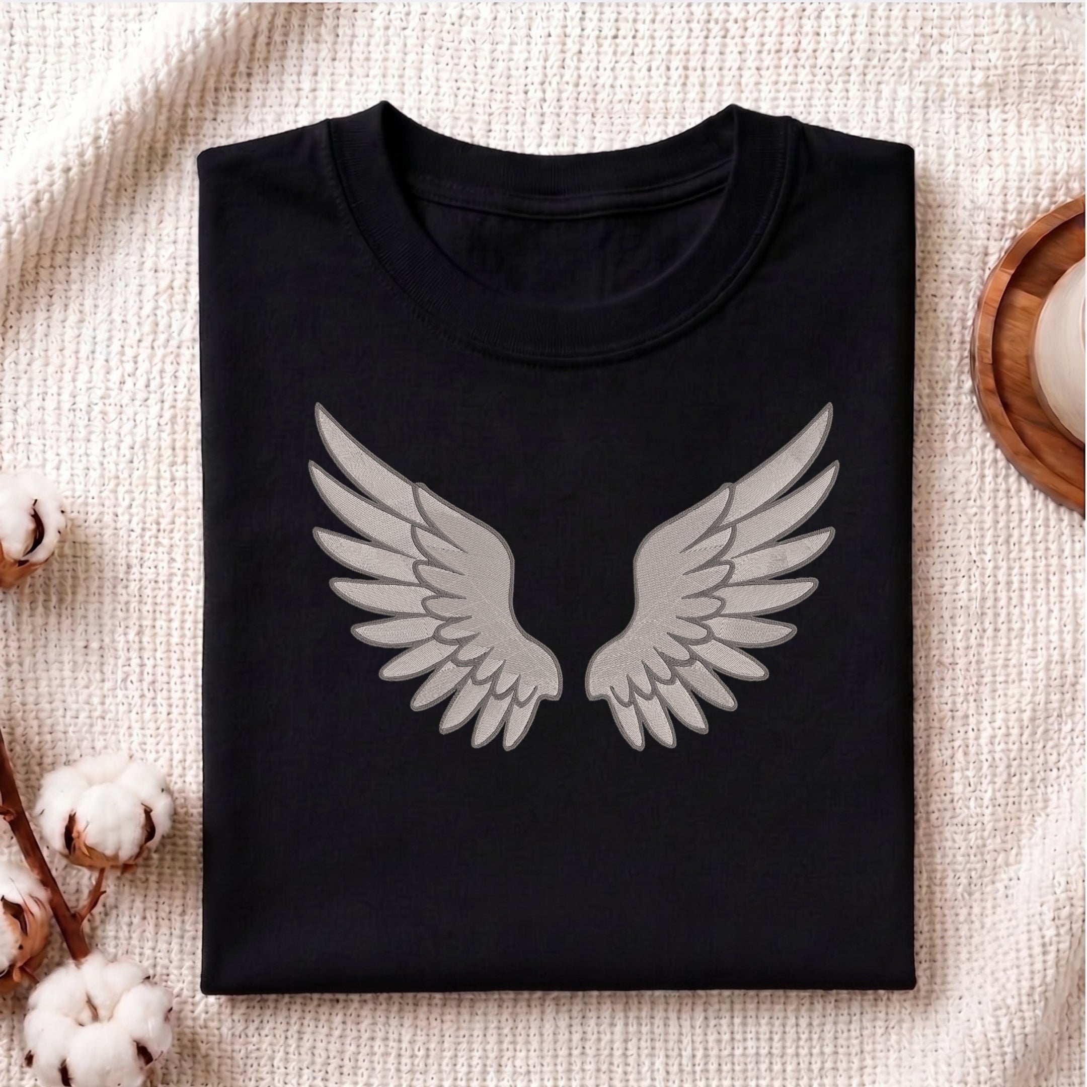 A folded black crewneck t-shirt with a gray angel wings graphic on the front, displayed on a textured beige fabric background with decorative cotton bolls and a wooden slice.