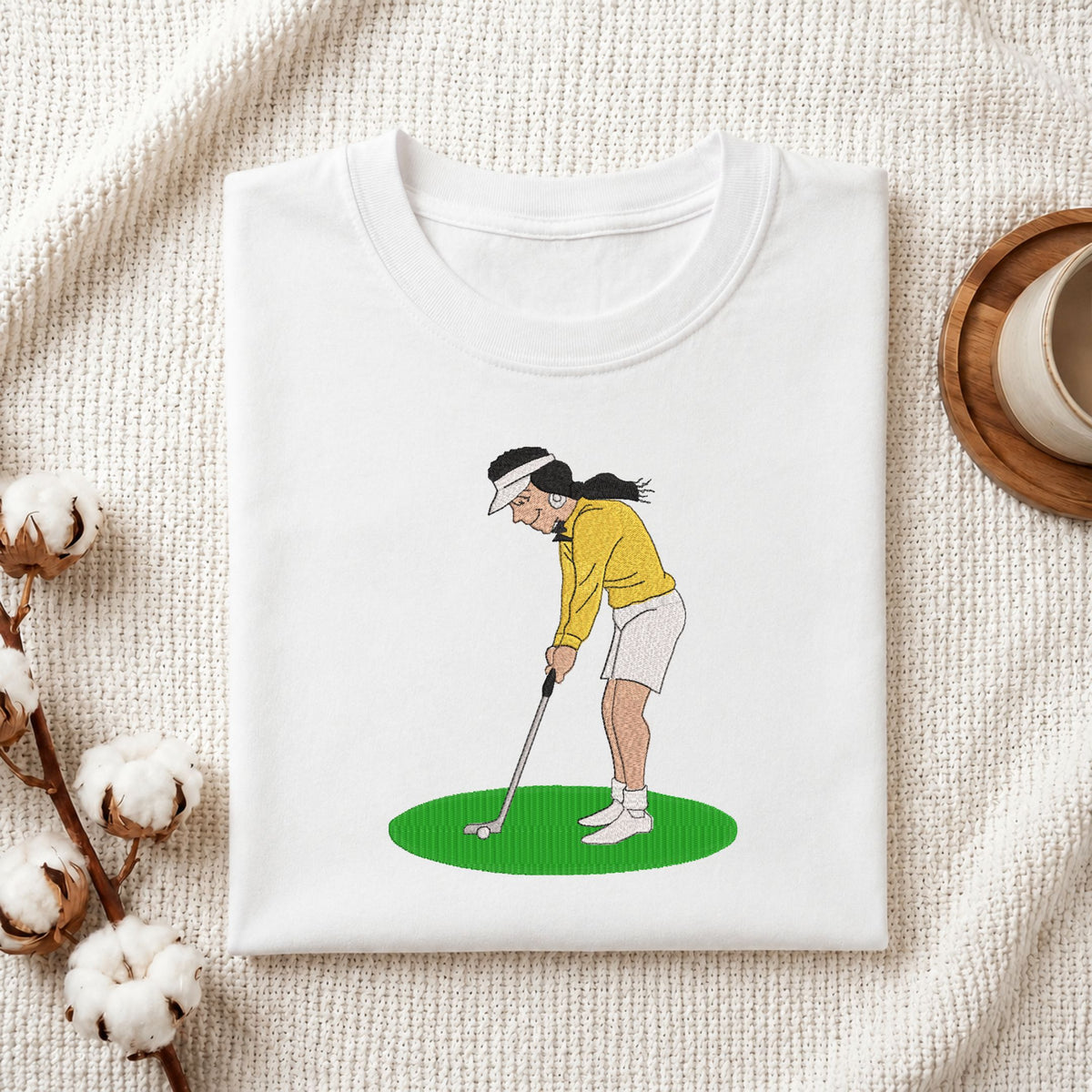  Folded white cotton t-shirt mockup featuring a woman playing golf machine embroidery design, styled with cotton stems and a wooden cup   