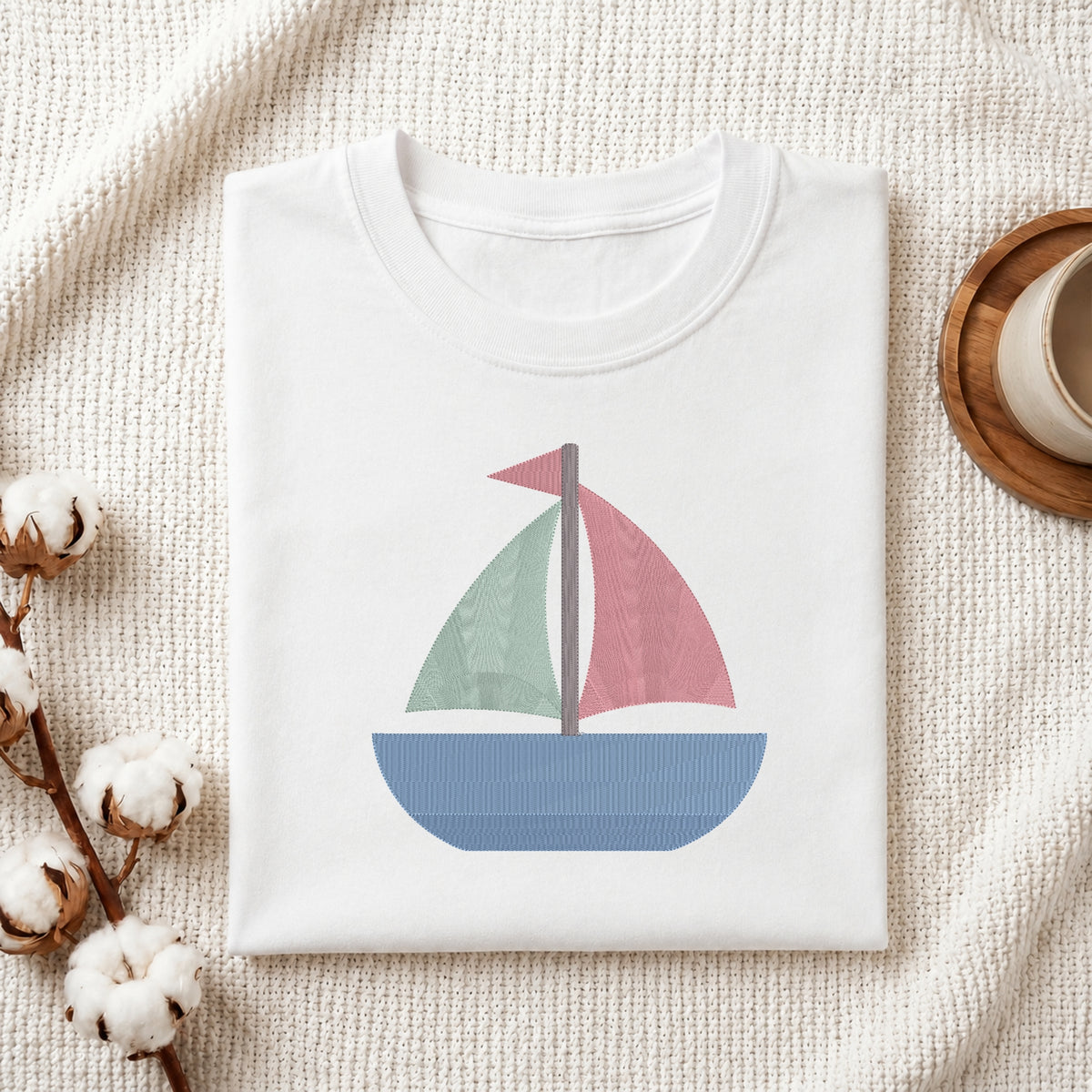 A neatly folded white crewneck t-shirt featuring a stitched colorful sailboat design, styled with cotton branches and a wooden coaster.
