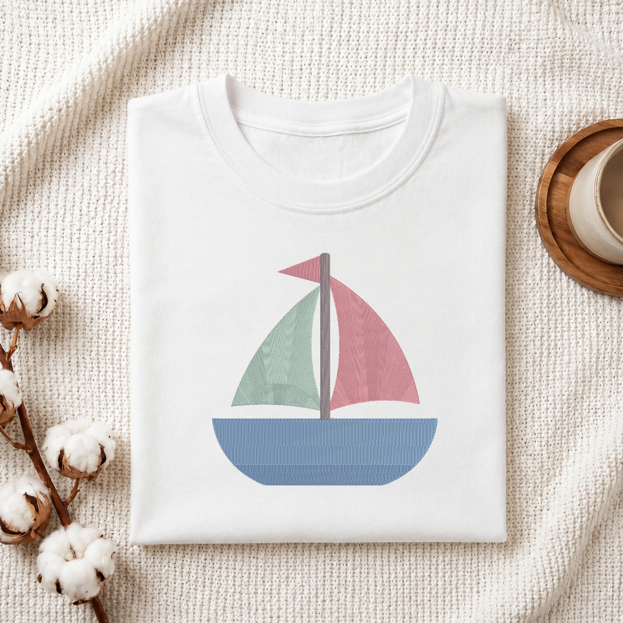 A neatly folded white crewneck t-shirt featuring a stitched colorful sailboat design, styled with cotton branches and a wooden coaster.