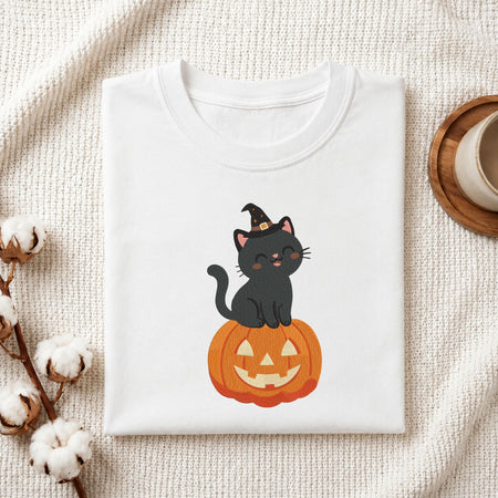 Folded white cotton t-shirt mockup featuring a cute black cat wearing a witch hat sitting on a jack-o-lantern pumpkin machine embroidery design, styled with cotton stems and a wooden cup