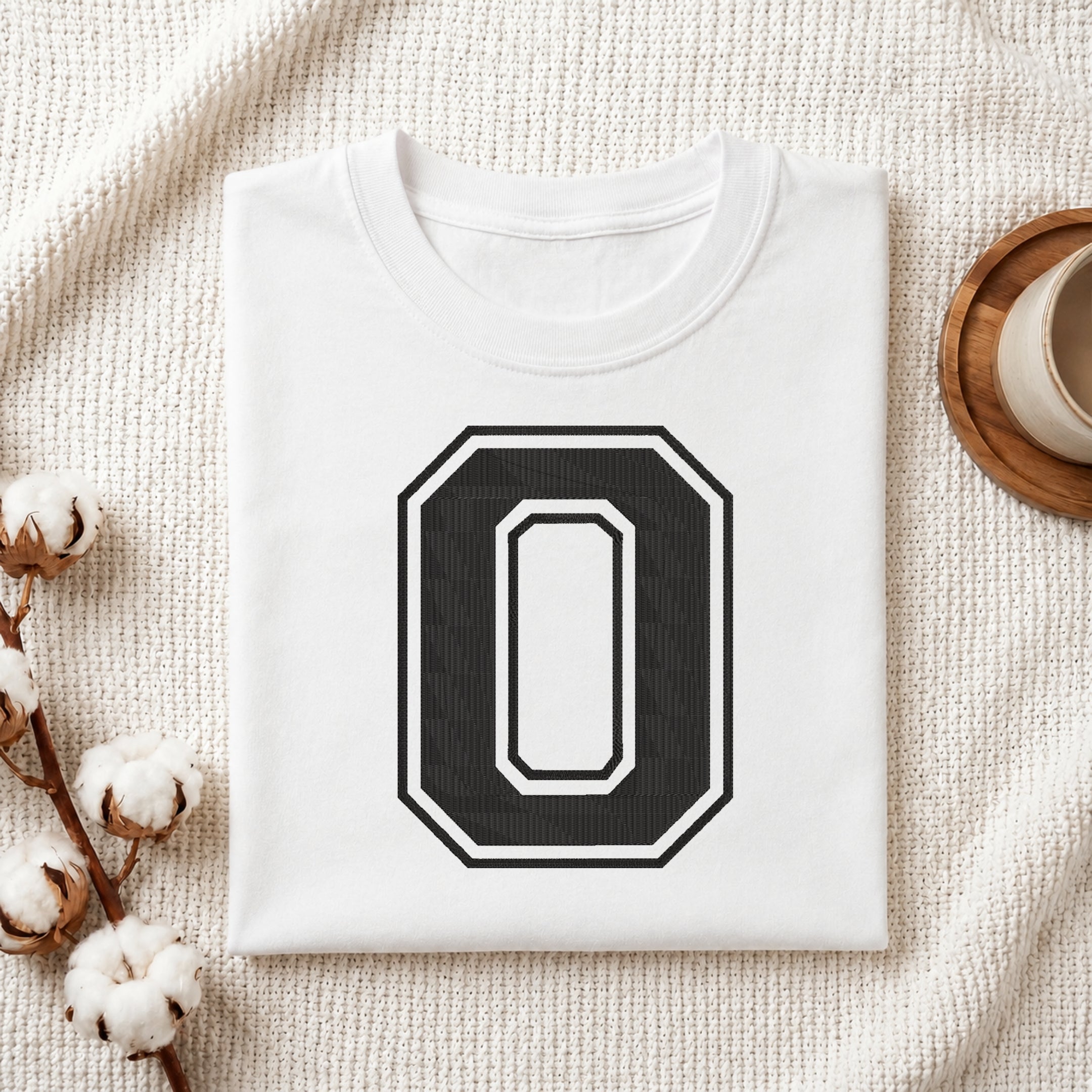 A folded white t-shirt with a large black and white number 0 graphic on the front, styled on a textured cream blanket with cotton bolls and a cup in the background.
