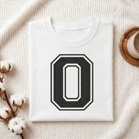A folded white t-shirt with a large black and white number 0 graphic on the front, styled on a textured cream blanket with cotton bolls and a cup in the background.