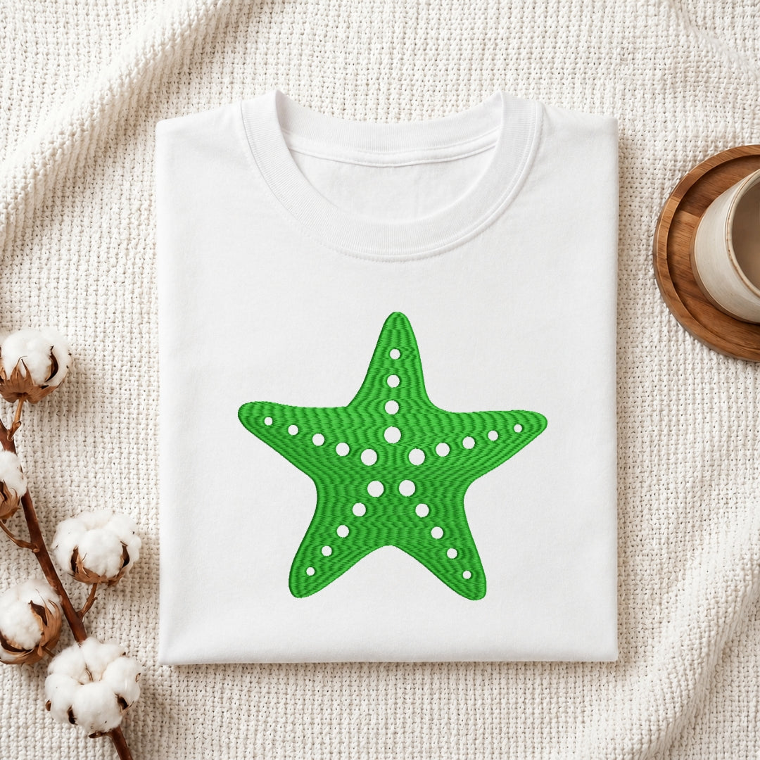 A folded white crewneck t-shirt featuring a bright green starfish graphic with white dots, styled on a textured cream fabric with cotton bolls and a wooden cup.