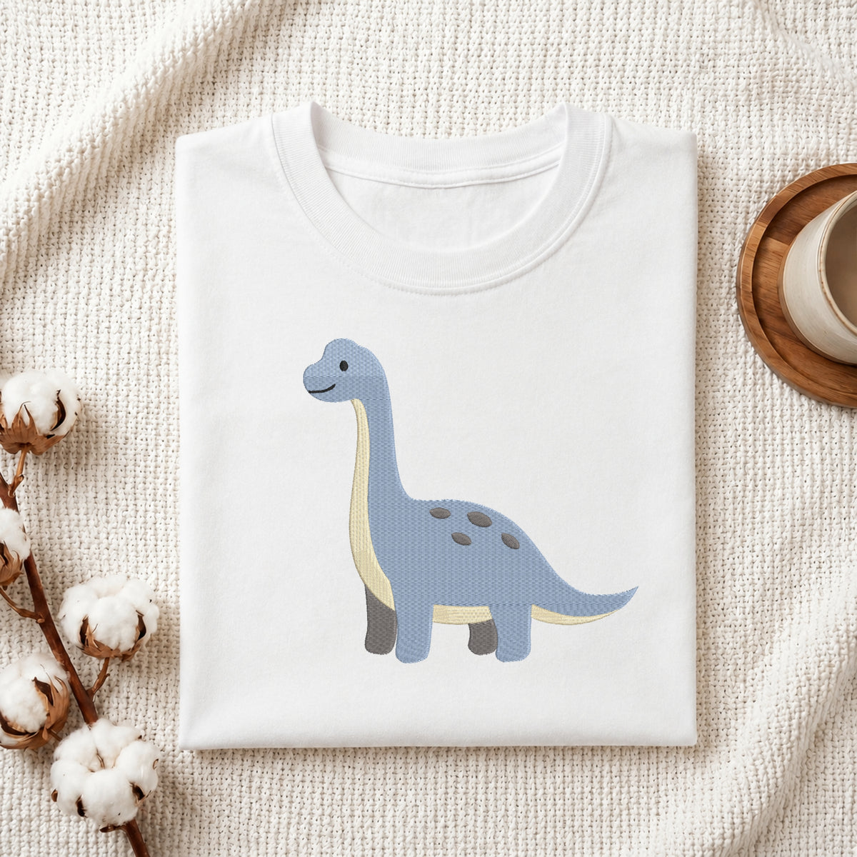 A folded white crewneck t-shirt featuring a cute light blue long-necked dinosaur with a cream belly, styled on a textured cream fabric with cotton bolls and a wooden cup in the background.