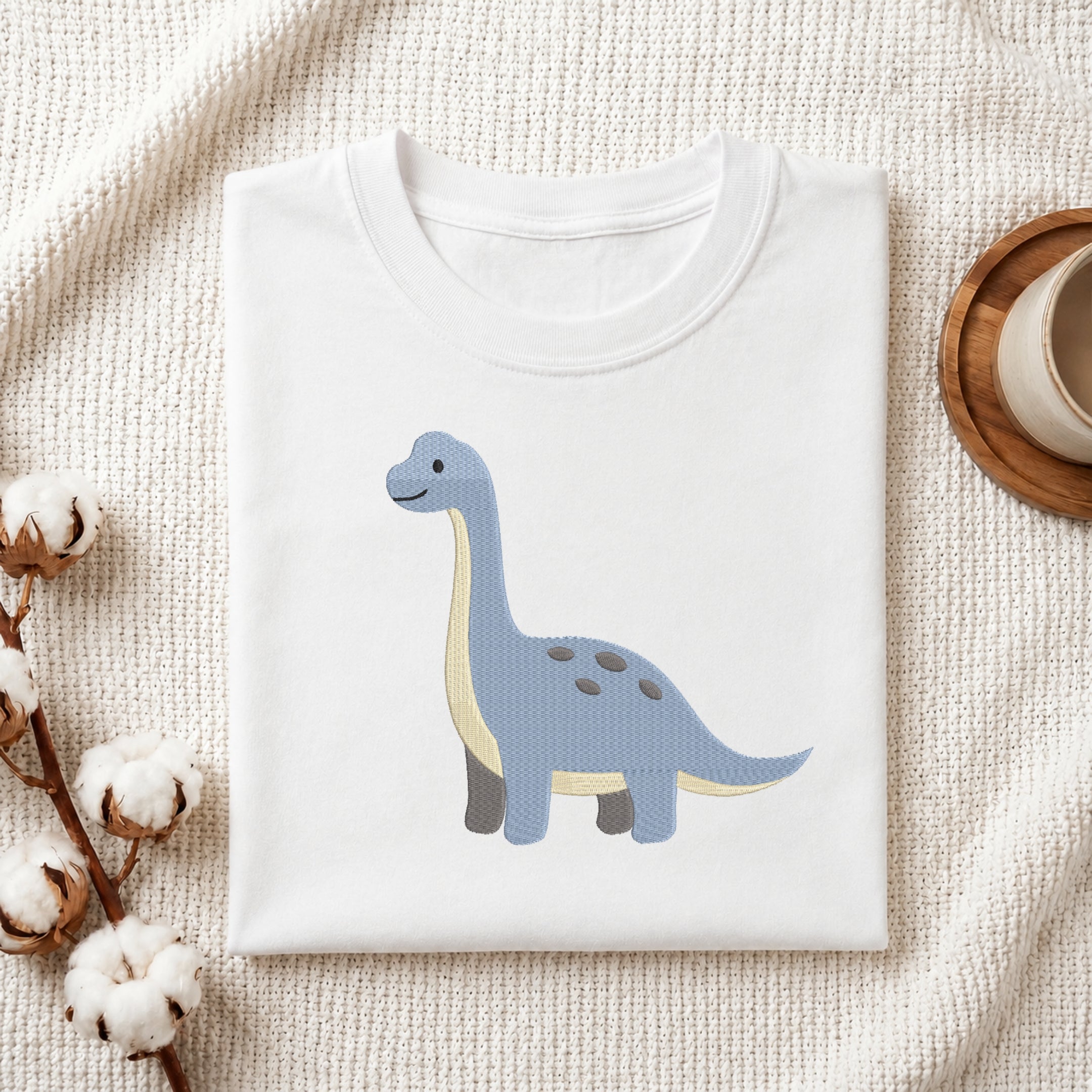A folded white crewneck t-shirt featuring a cute light blue long-necked dinosaur with a cream belly, styled on a textured cream fabric with cotton bolls and a wooden cup in the background.