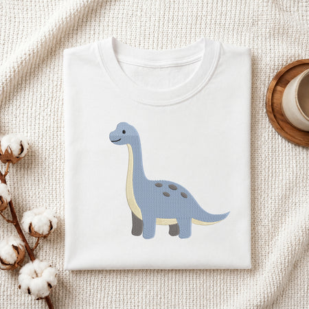 A folded white crewneck t-shirt featuring a cute light blue long-necked dinosaur with a cream belly, styled on a textured cream fabric with cotton bolls and a wooden cup in the background.